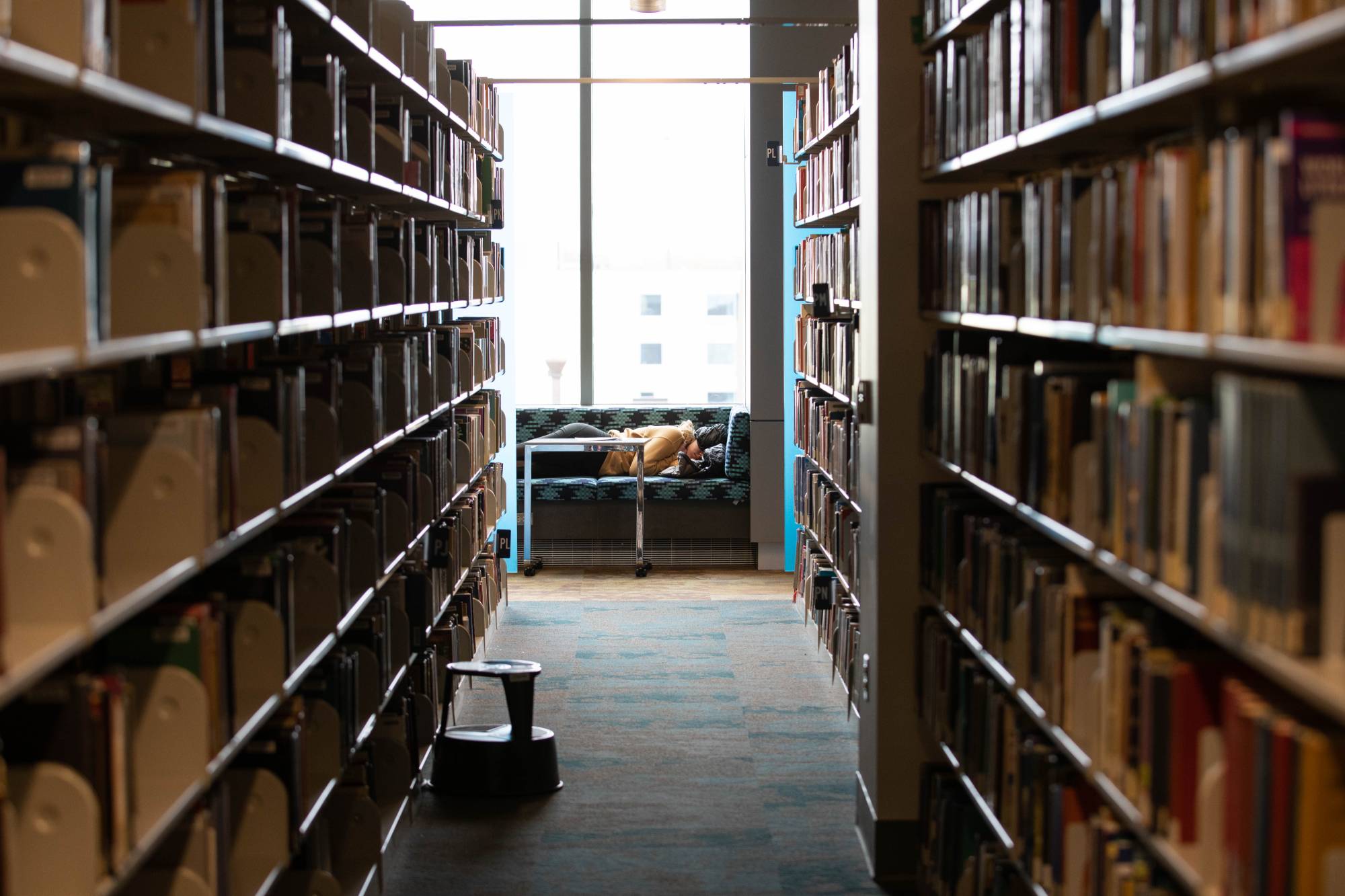 Person sleeping on library seating at the end of bookshelf row